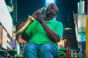 En la imagen, un hombre vestido con una camisa verde y pantalones de mezclilla está sentado, interpretando la saxofón en un lugar bullicioso. Las pantallas gigantes y los letreros iluminados en el fondo sugieren que se encuentra en un área céntrica, quizás Times Square. Al igual que la música del saxofón atrae a la gente que pasa por el lugar, BaoLiba se dedica a conectar a los influencers y las marcas en Colombia. Facilita la interacción entre ellos, creando oportunidades que, como la melodía en medio de la agitación de la ciudad, logran destacar y generar un impacto.