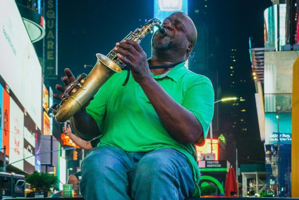 En la imagen, un hombre vestido con una camisa verde y pantalones de mezclilla está sentado, interpretando la saxofón en un lugar bullicioso. Las pantallas gigantes y los letreros iluminados en el fondo sugieren que se encuentra en un área céntrica, quizás Times Square. Al igual que la música del saxofón atrae a la gente que pasa por el lugar, BaoLiba se dedica a conectar a los influencers y las marcas en Colombia. Facilita la interacción entre ellos, creando oportunidades que, como la melodía en medio de la agitación de la ciudad, logran destacar y generar un impacto.