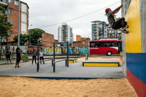 En la imagen, un individuo está practicando un movimiento de escalada en una pared colorida, mientras que varios espectadores lo observan en un espacio abierto urbano. Hay edificios en el fondo, un autobús rojo y murales de graffiti en el entorno. Las tendencias del marketing en redes sociales en Colombia para 2025, similar a este escena dinámica, se centrarán en la interacción y la autenticidad. Las marcas buscarán conectarse con la audiencia a través de contenido real, como la práctica deportiva en entornos cotidianos, y fomentar la participación activa de los usuarios, así como aprovechar los ambientes locales y auténticos para resonar con la cultura y la vida de los colombianos.